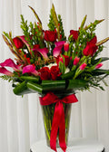 Bouquet of red roses, pink calla lilies and tulips in a clear vase with a red ribbon against a white curtain background